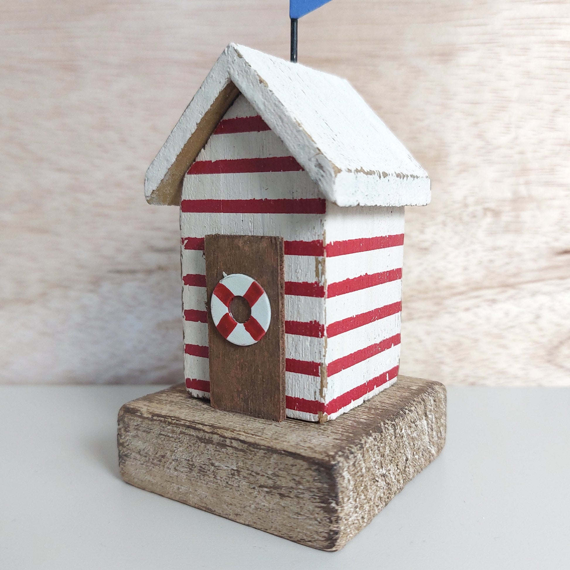 A small decorative beach hut figurine with red and white horizontal stripes, a weathered brown door with a red-and-white life preserver detail, and a rustic wooden base, photographed against a light neutral background.