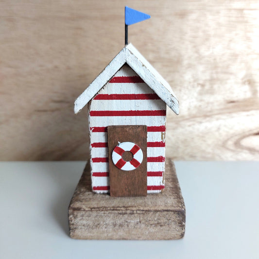 A small decorative beach hut figurine with red and white horizontal stripes, a white roof with a blue flag, a brown door with a red-and-white life preserver, and a rustic wooden base.