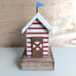 A small decorative beach hut figurine with red and white horizontal stripes, a white roof with a blue flag, a brown door with a red-and-white life preserver, and a rustic wooden base.