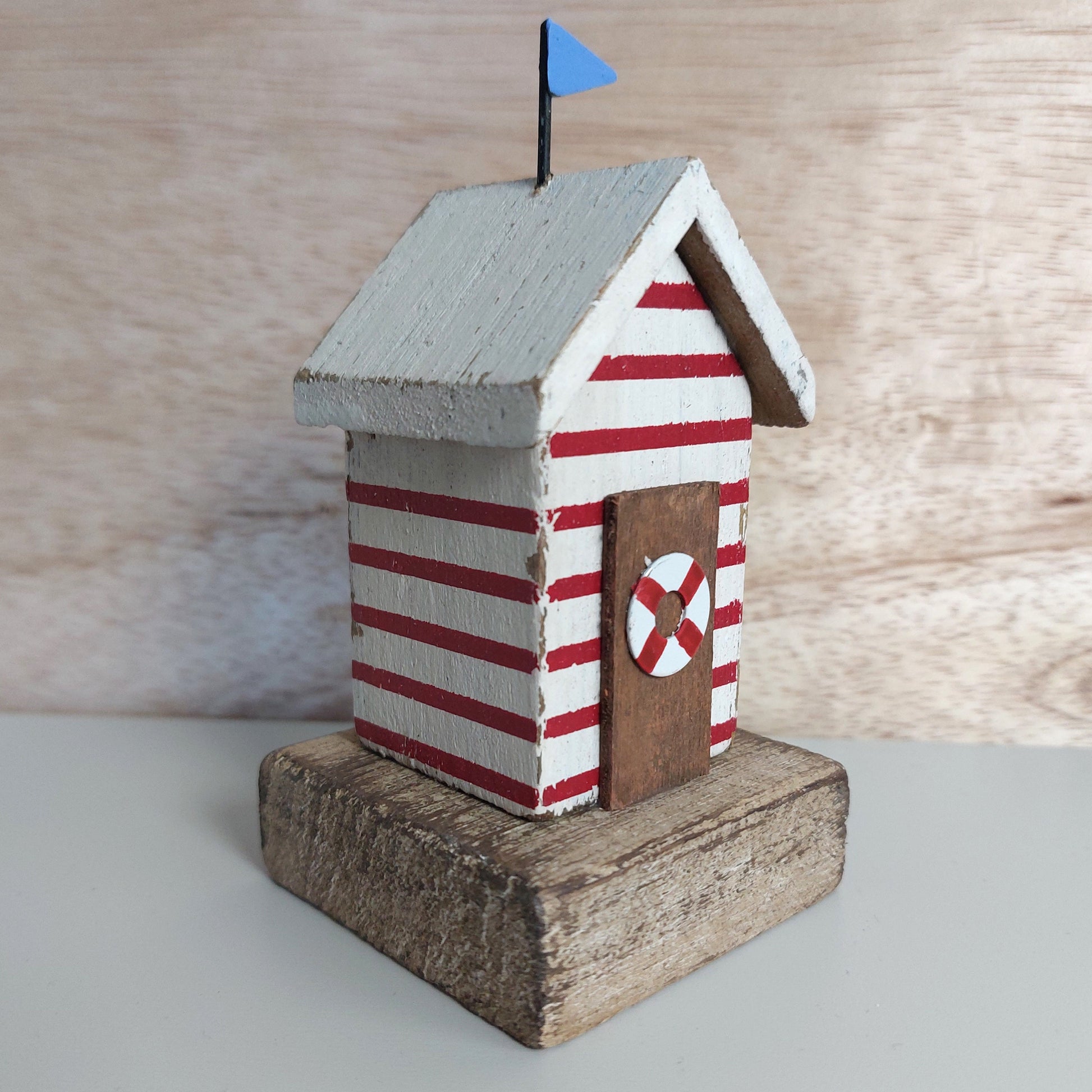 A small decorative beach hut ornament with red and white horizontal stripes, a brown door, a small blue flag on top, and a rustic wooden base, photographed against a light wood background.