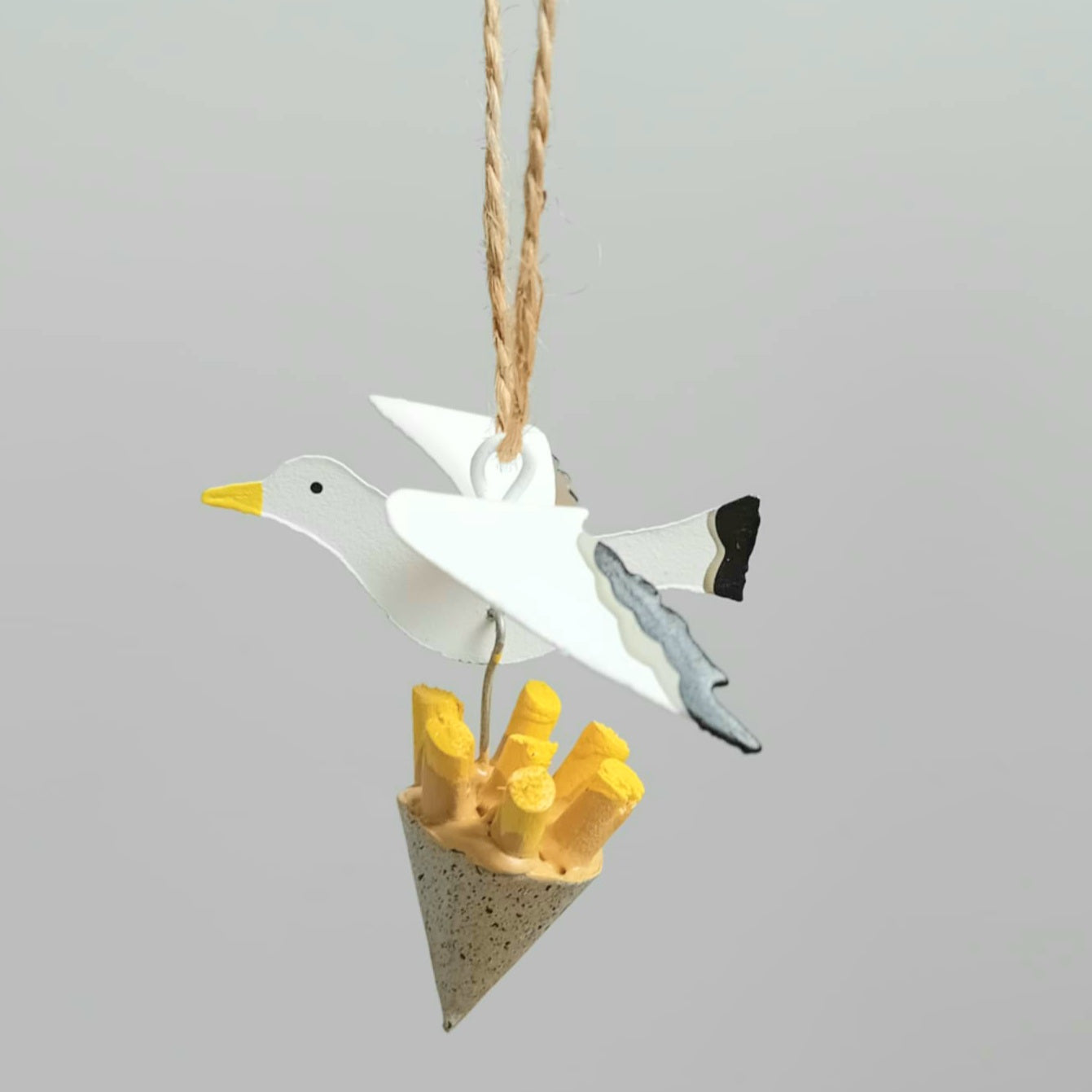 Decorative ornament of a seagull carrying some chips in a packet, hanging from a string against a light gray background.