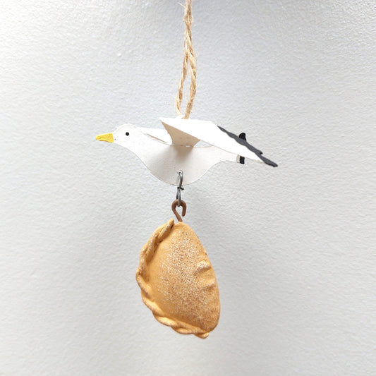 A metal seagull carrying a pasty away hanging from a jute string against a grey background
