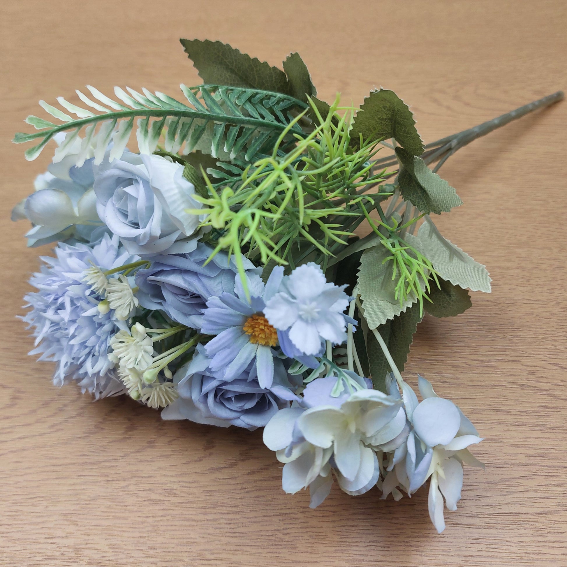 An artificial floral bouquet with light blue and white faux flowers, green leaves, and fern-like stems on a light wood surface.