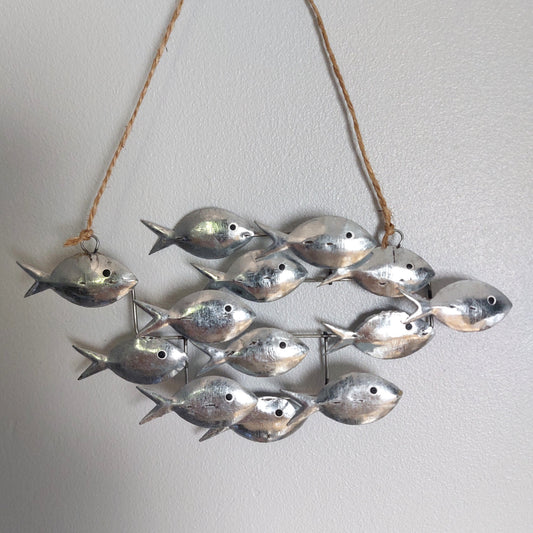 A hanging wall decor piece made of multiple small silver fish attached to twine, shown against a plain light gray wall.