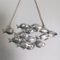 A hanging wall decor piece made of multiple small silver fish attached to twine, shown against a plain light gray wall.