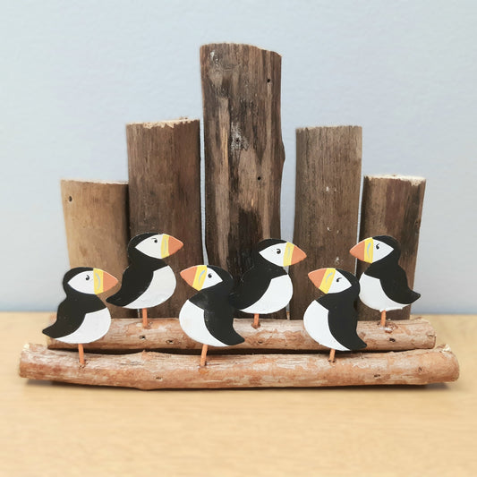 A model of 6 metal puffins with some wooden groynes and driftwood against a grey background