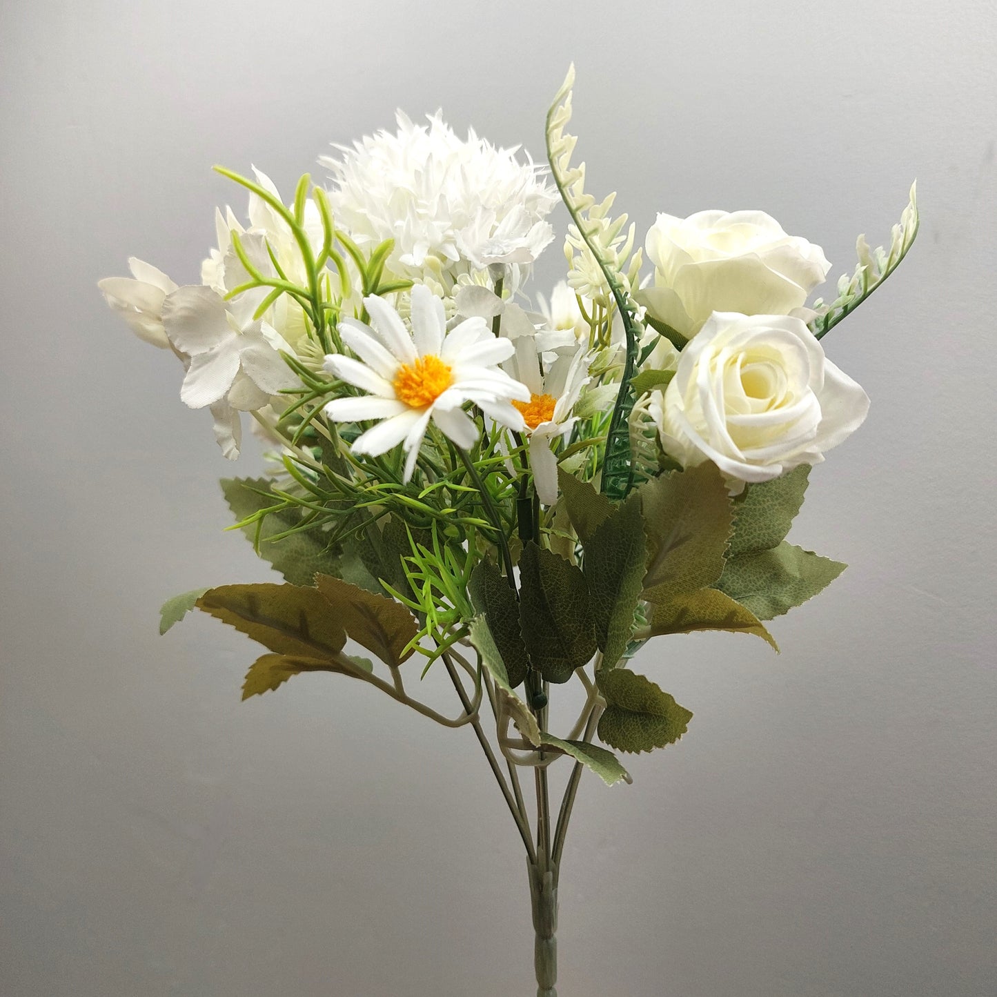 An artificial flower bouquet featuring white roses, white daisies with yellow centers, and green foliage, set against a plain light gray background.