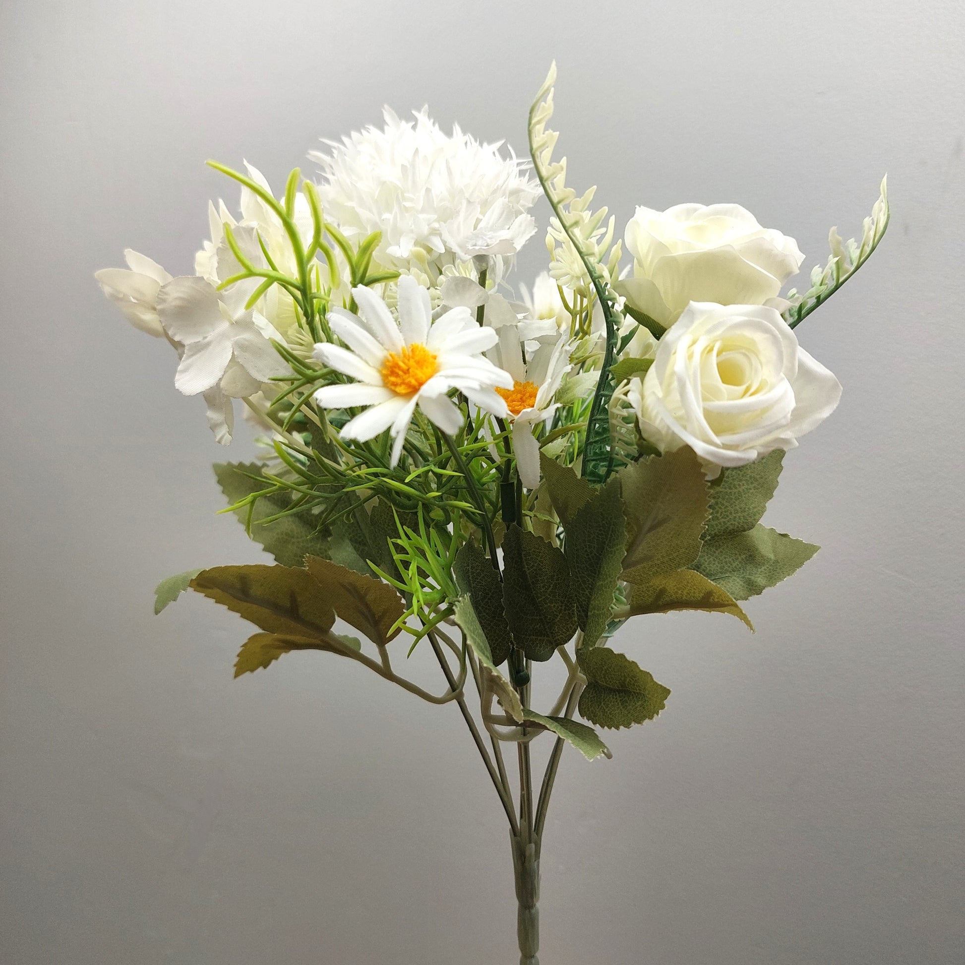 An artificial flower bouquet featuring white roses, white daisies with yellow centers, and green foliage, set against a plain light gray background.
