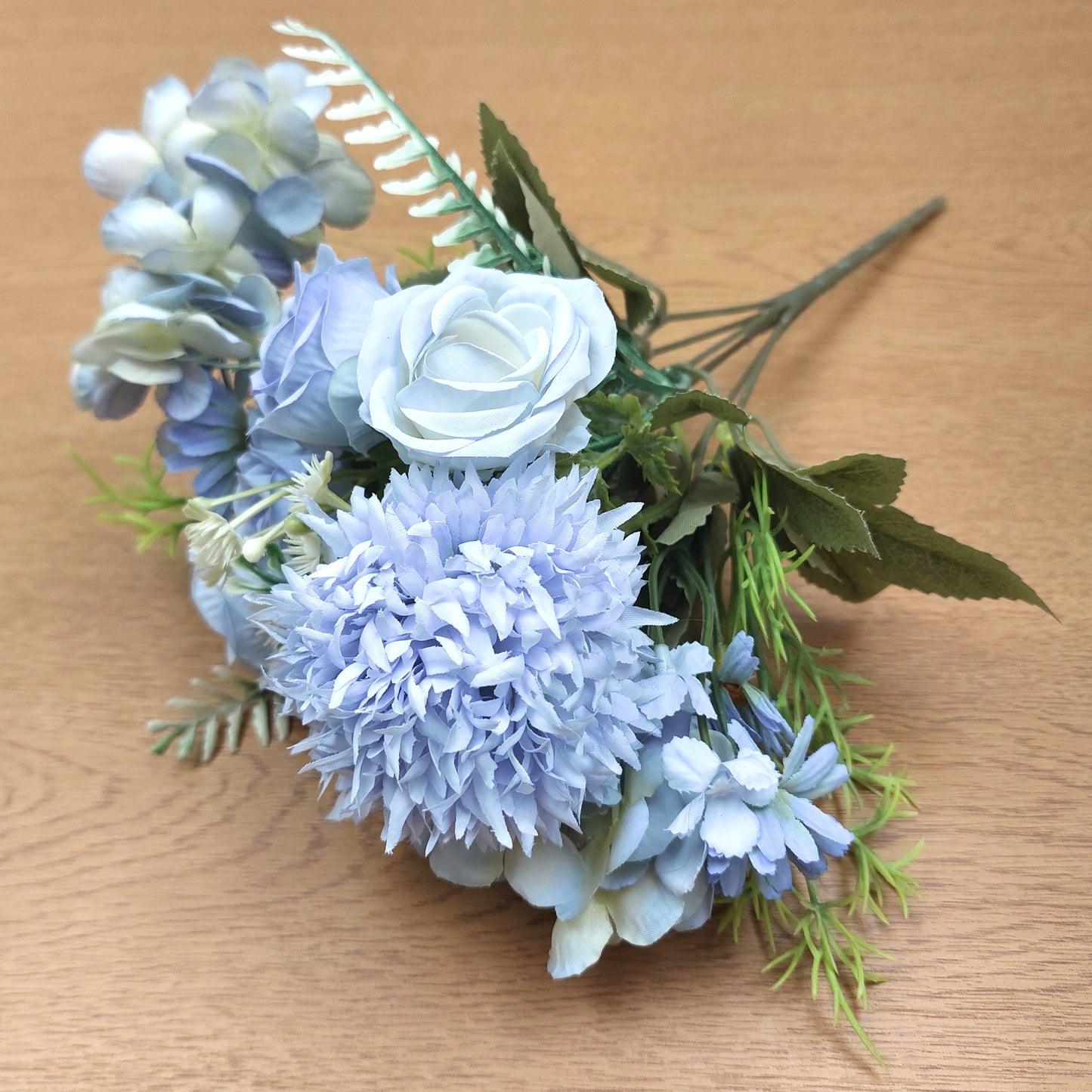An artificial floral bouquet featuring light blue hydrangeas, a pale blue rose, and green leaves, displayed on a light brown wooden surface.