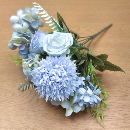 An artificial floral bouquet featuring light blue hydrangeas, a pale blue rose, and green leaves, displayed on a light brown wooden surface.
