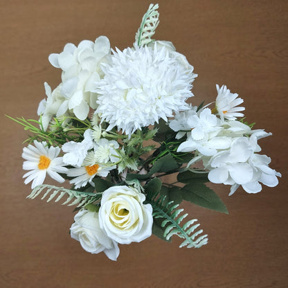 Ivory artificial flower bouquet with roses, daisies, and green foliage on a brown background.
