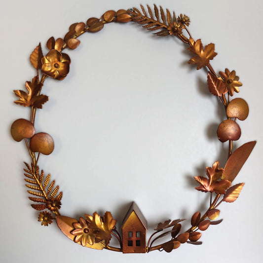 A whole tarnished bronze style metal wreath with foliage, flowers and a cottage against a pale background
