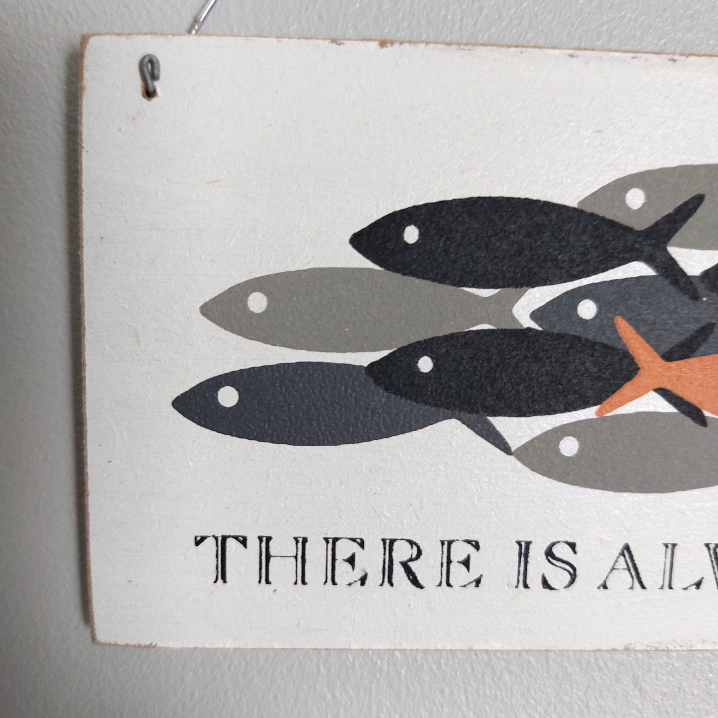 A rectangular white decorative sign with a printed illustration of several fish in black, gray, and orange, plus the partial text 'THERE IS ALW' at the bottom, hanging against a light gray wall.