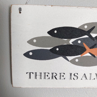 A rectangular white decorative sign with a printed illustration of several fish in black, gray, and orange, plus the partial text 'THERE IS ALW' at the bottom, hanging against a light gray wall.