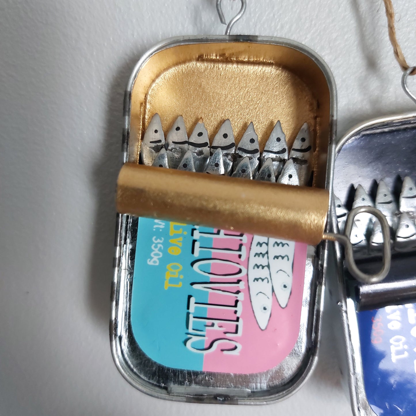 A handmade tinned fish ornament or wall hanging made from an open metal tinned fish can, decorated with small silver anchovy fish, and a colorful label on the bottom half.