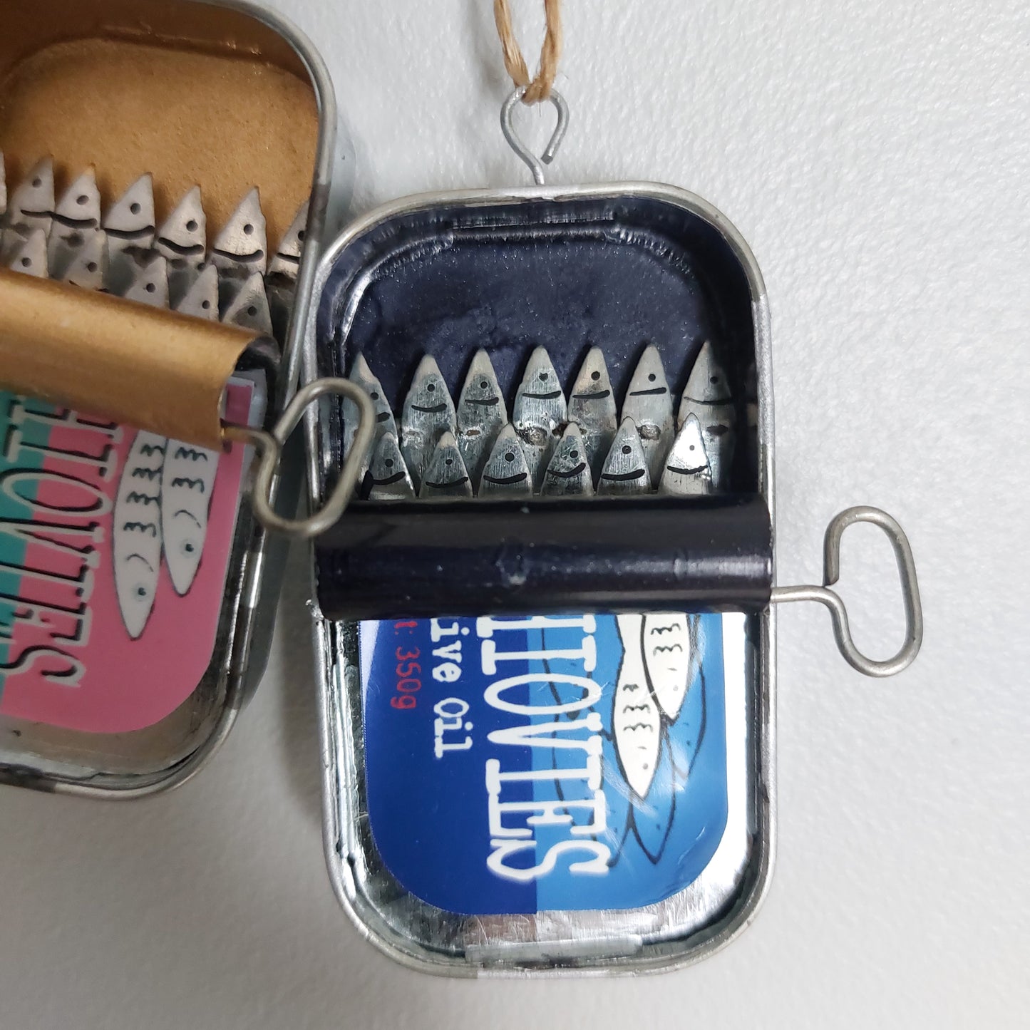 A novelty wall hanging or ornament shaped like an open sardine or anchovy tin with metal anchovy fish sticking out, shown against a plain light wall. A second similar piece is partially visible next to it as a set of two.