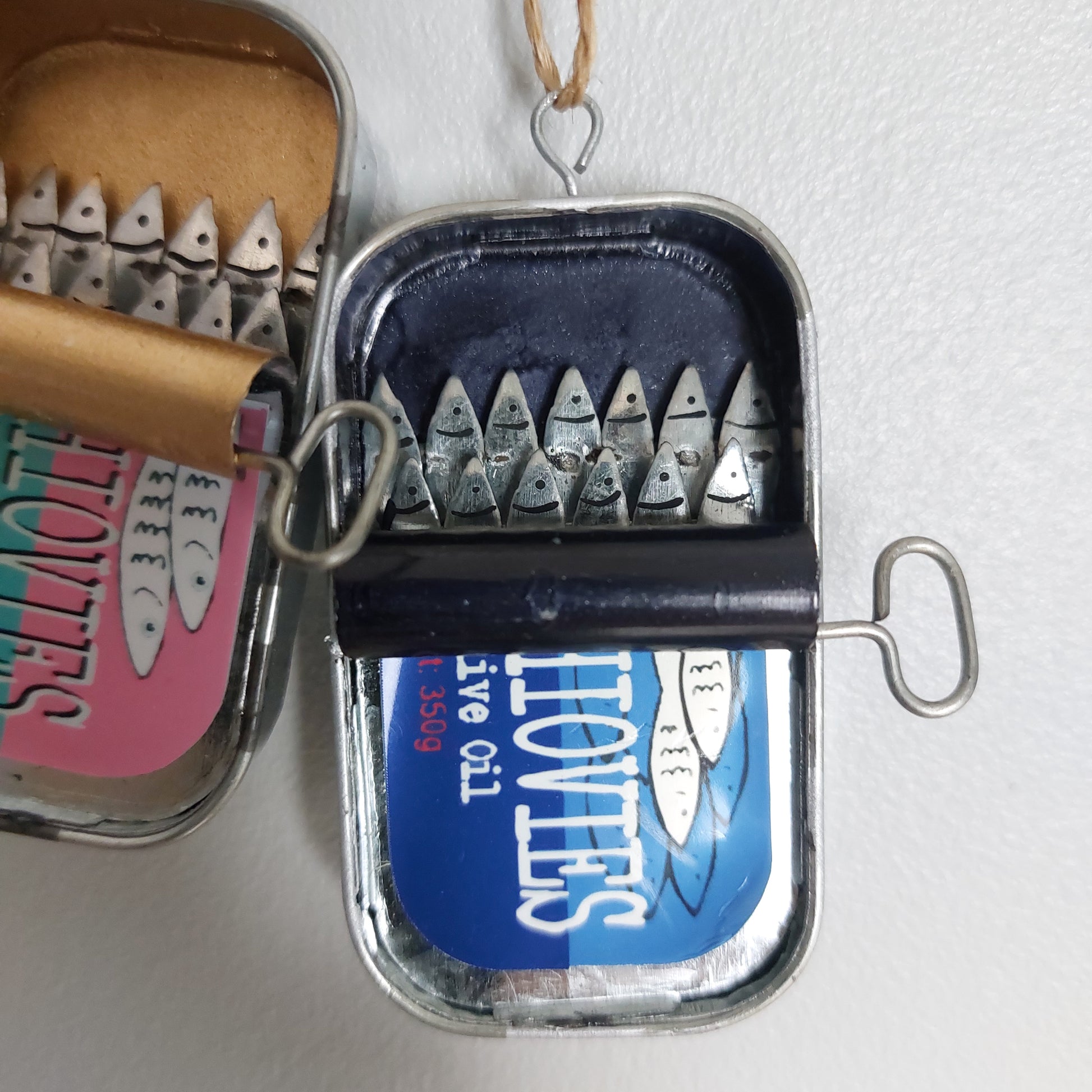 A novelty wall hanging or ornament shaped like an open sardine or anchovy tin with metal anchovy fish sticking out, shown against a plain light wall. A second similar piece is partially visible next to it as a set of two.