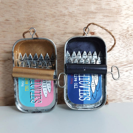 Two ornamental tins with metal anchovies peeking out, with colorful Aqua, Pink and Blue labels, displayed on a light surface against a wood background.