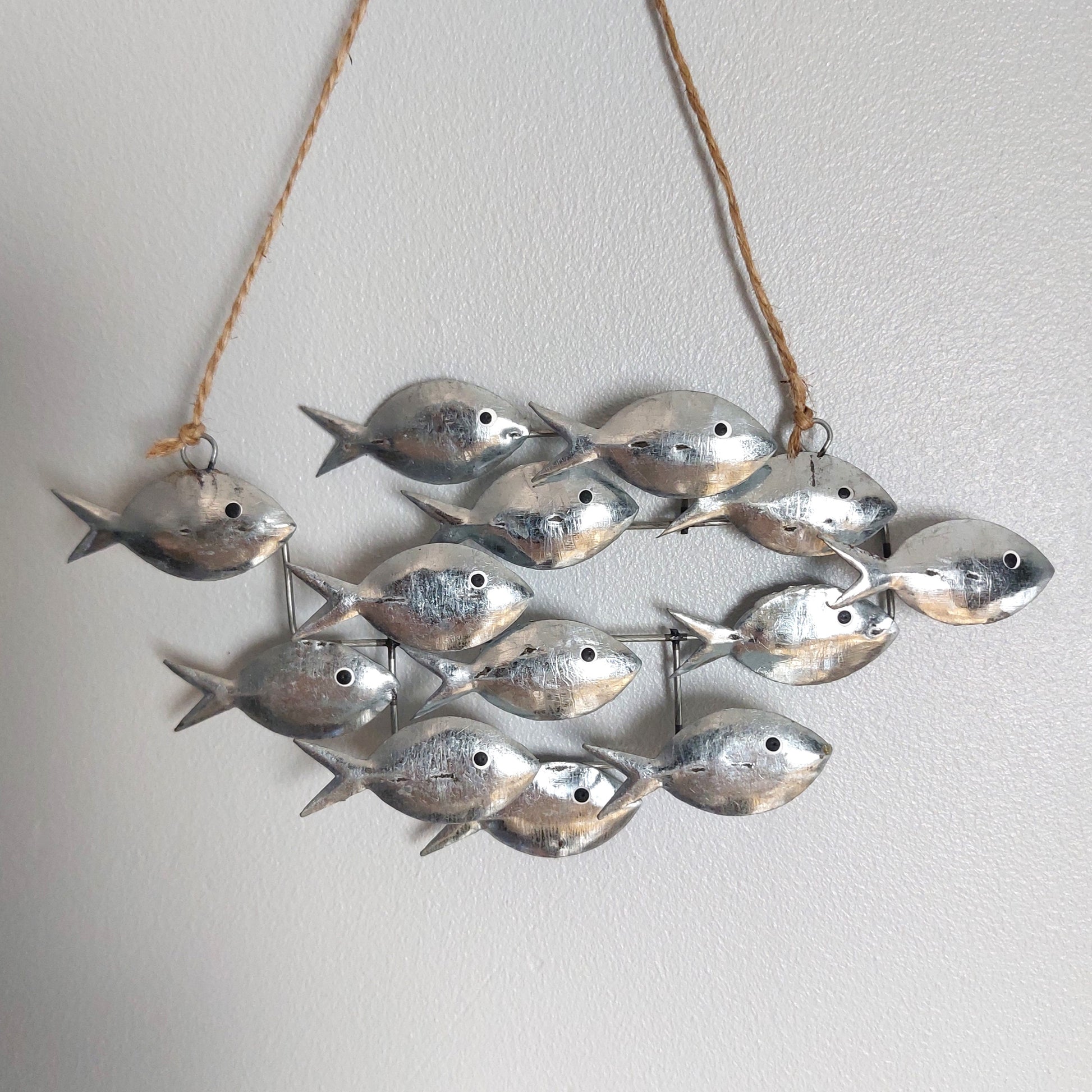 A decorative hanging wall piece made of multiple small metallic fish charms suspended from a natural twine string against a plain light wall.