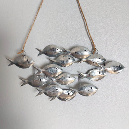 A decorative hanging wall piece made of multiple small metallic fish charms suspended from a natural twine string against a plain light wall.