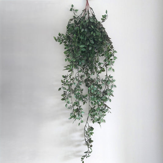A hanging artificial green leafy ruscus vine with trailing stems and small oval leaves, suspended against a plain light gray wall.