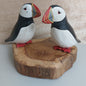 Two hand carved and painted puffin figurines on a wooden base against a neutral background