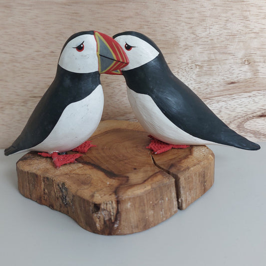 A pair of hand carved and painted wooden Puffins mounted on a split wood slice, against a pale background.