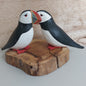 A pair of hand carved and painted wooden Puffins mounted on a split wood slice, against a pale background.