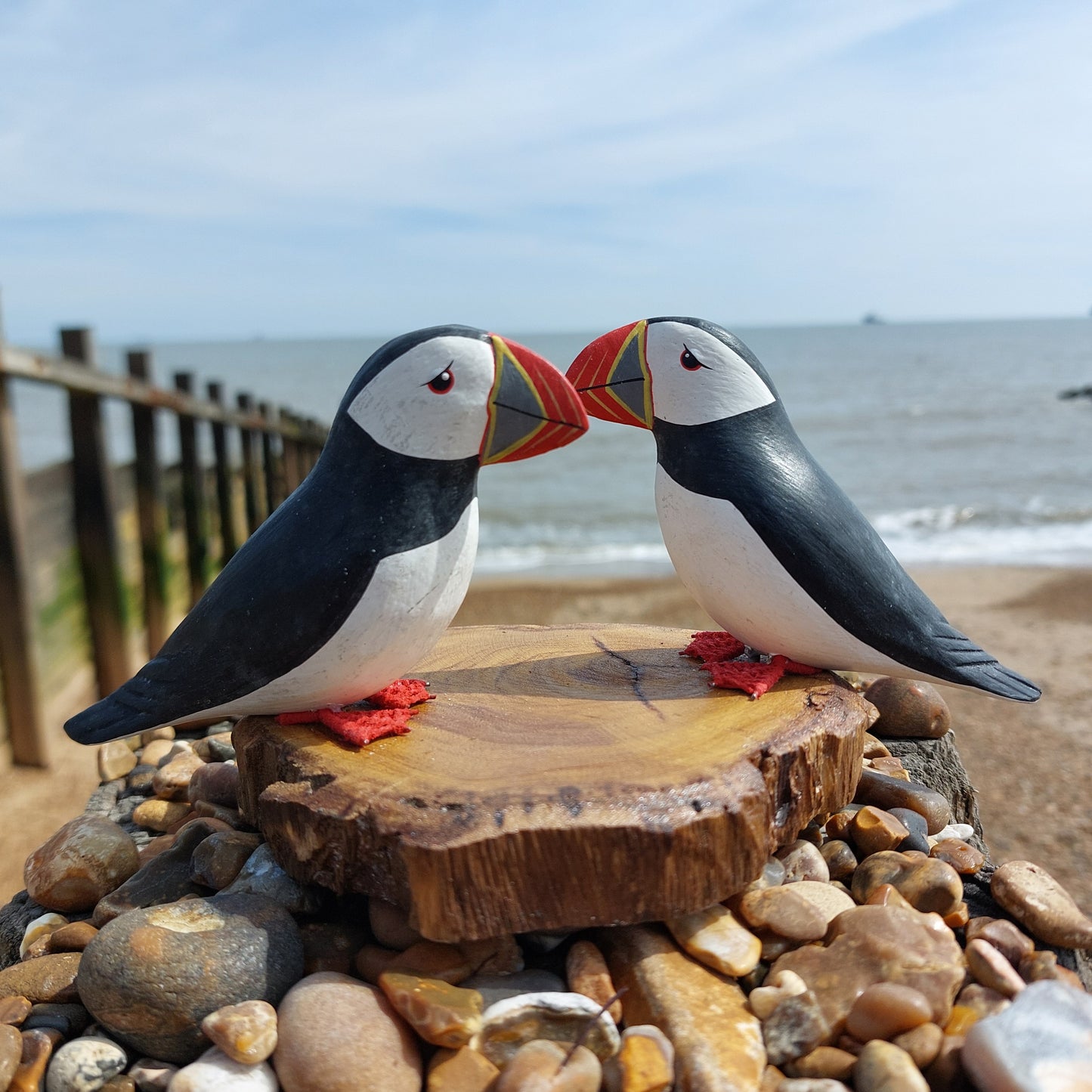 Two wooden hand carved and painted decorative puffins on a wooden platform with a beach and ocean background