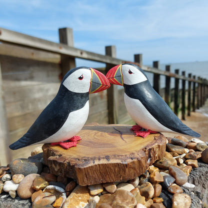 Two wooden puffin figures mounted on a wooden base on a wooden groyne by the sea.
