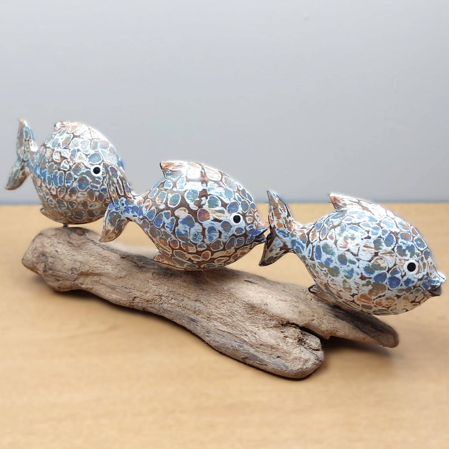 Three carved wooden fish with mosaic patterns on a piece of driftwood against a neutral background