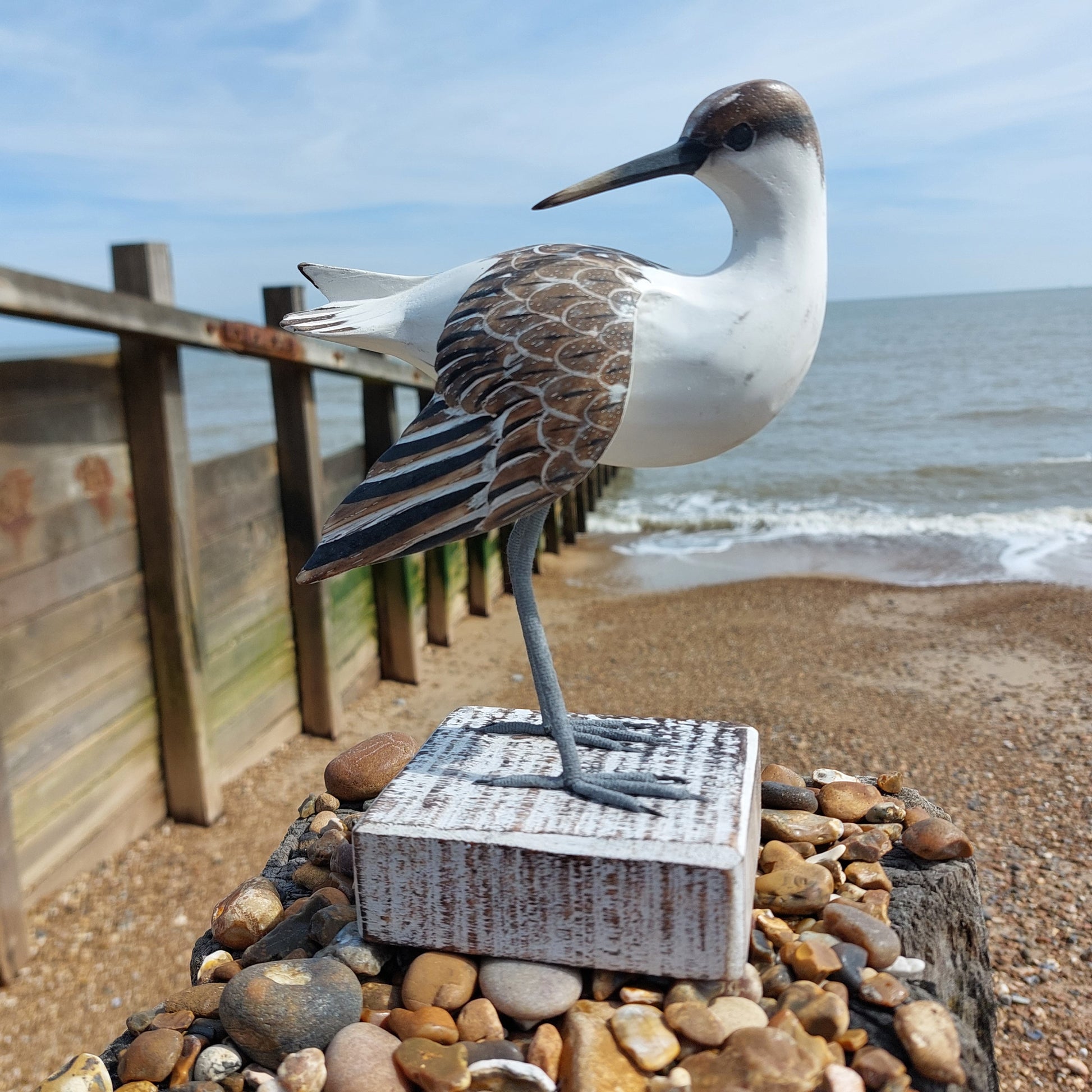 Decorative wooden Godwit bird sculpture on pebbles by a wooden railing with ocean view