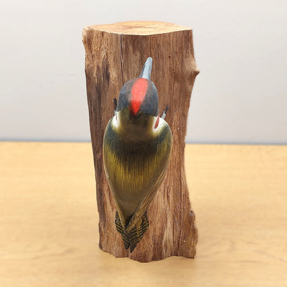 A wooden ornament from Bali of a Green Woodpecker clinging to a wooden log against a wooden and pale background.