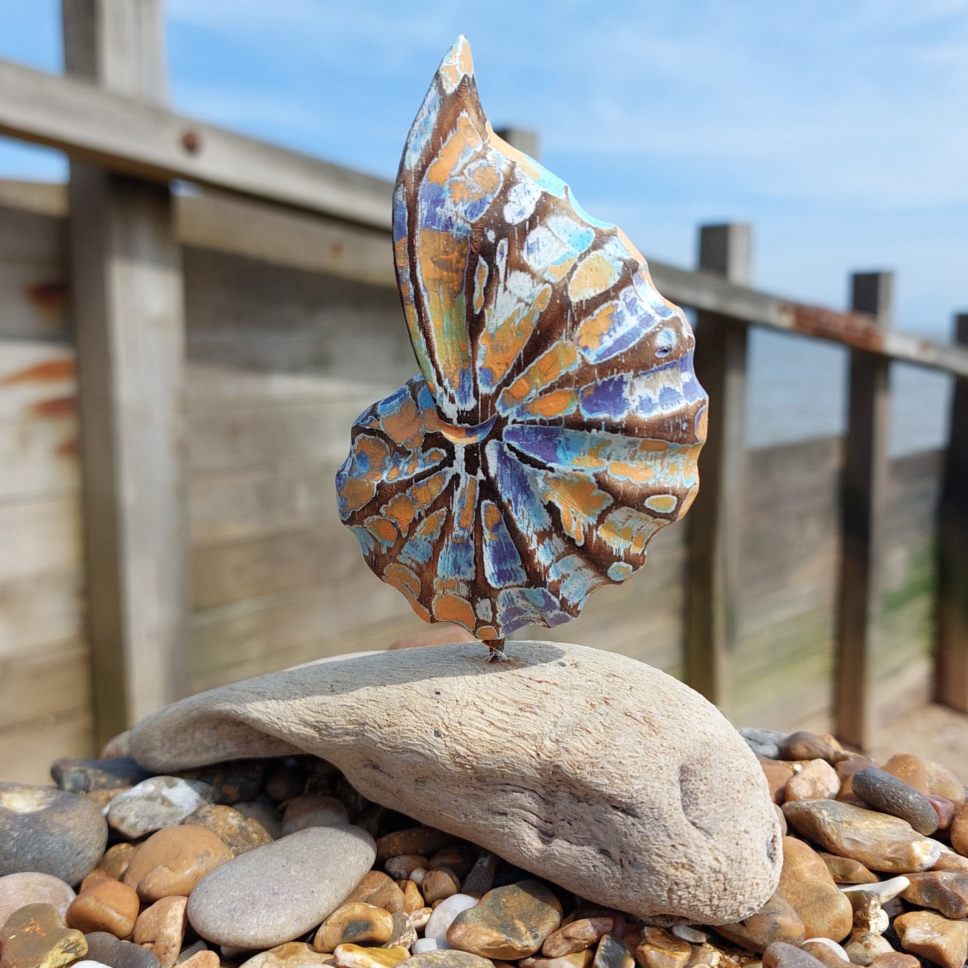 Decorative shell sculptures on a wooden driftwood piece with a wooden fence and blue sky in the background