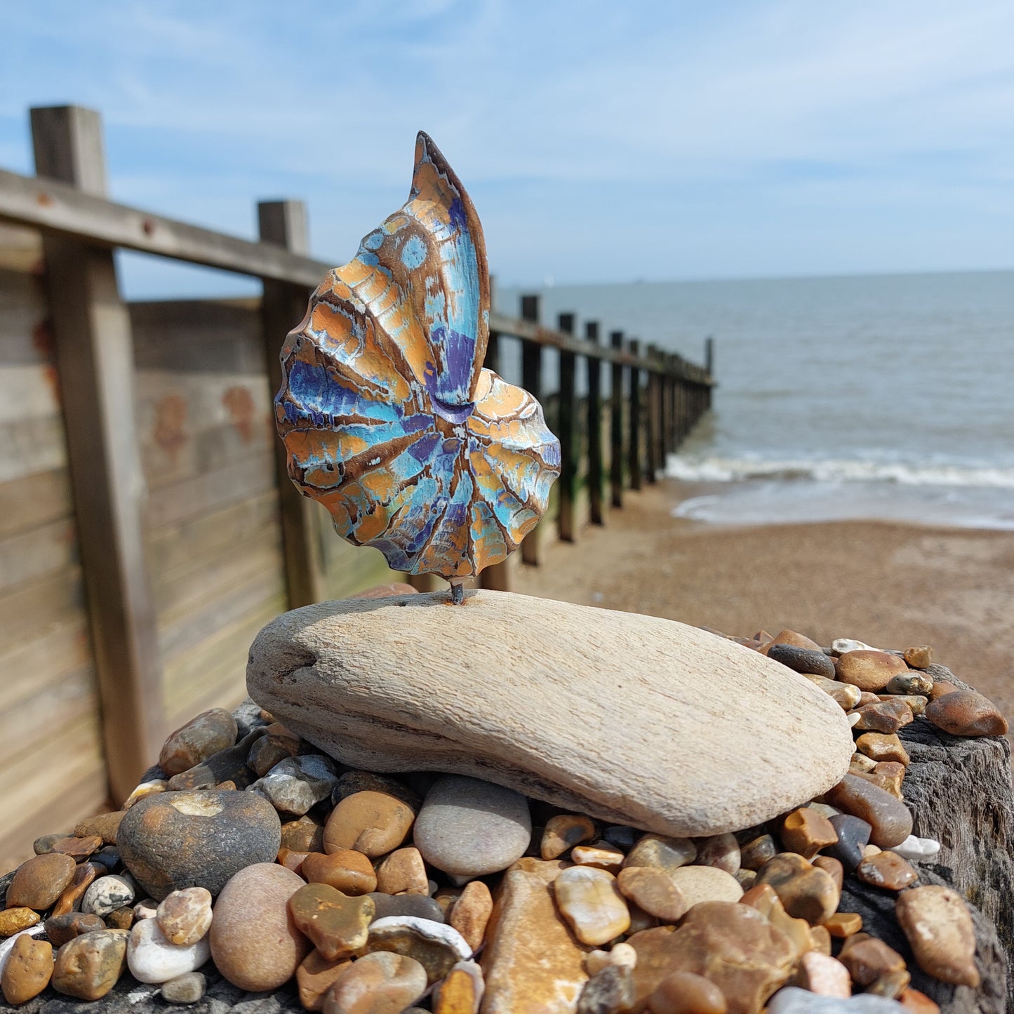 Decorative wooden shell ornament mounted on a piece of driftwood on some pebbles on a groyne with a background of the sea and beach