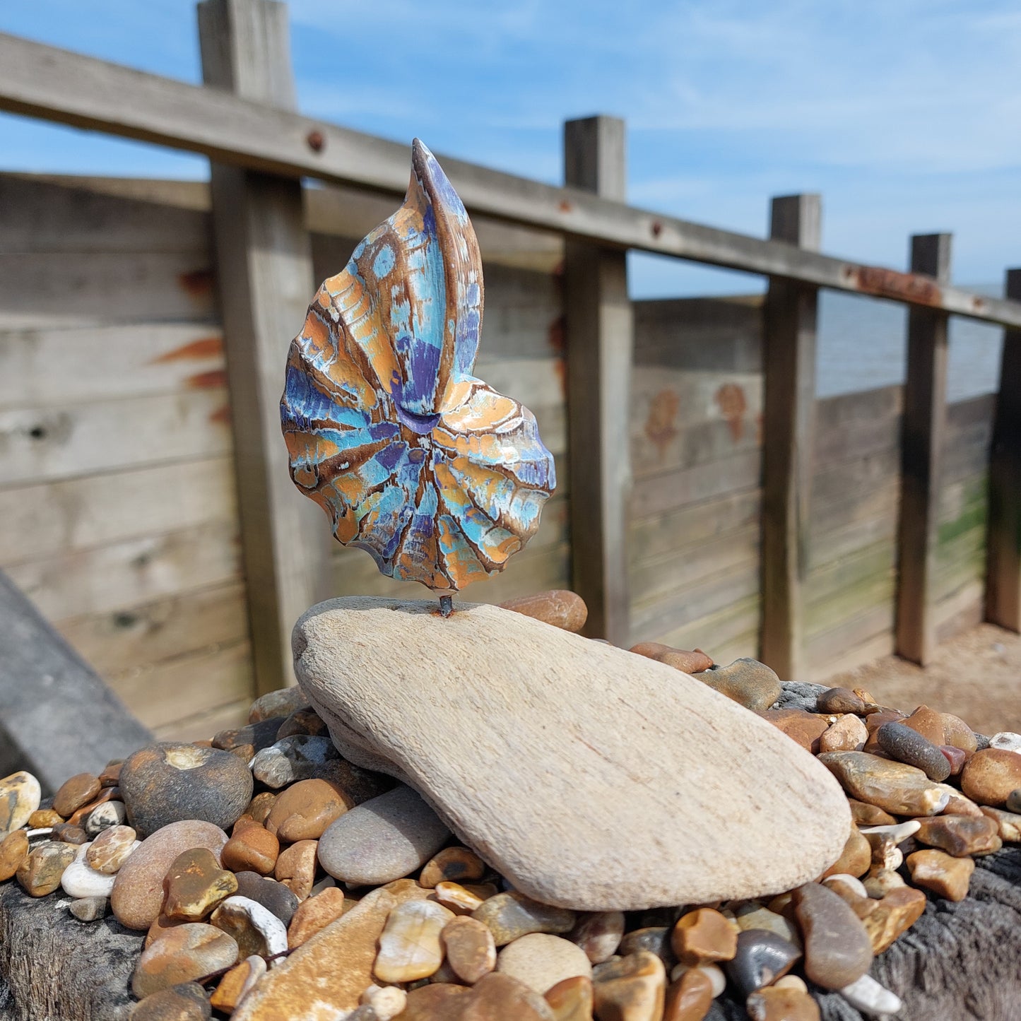 Decorative shell on a wooden groyne with some pebbles and blue sky in the background