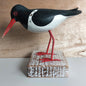 Hand carved Wooden Model of an Oystercatcher bird with a red beak and legs on a wooden block against a wooden background