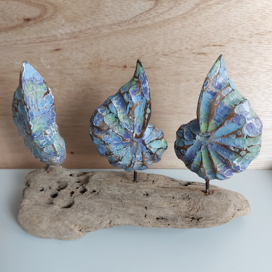 Three blue, green and brown hand carved wooden shells attached to a piece of driftwood on a wooden surface.